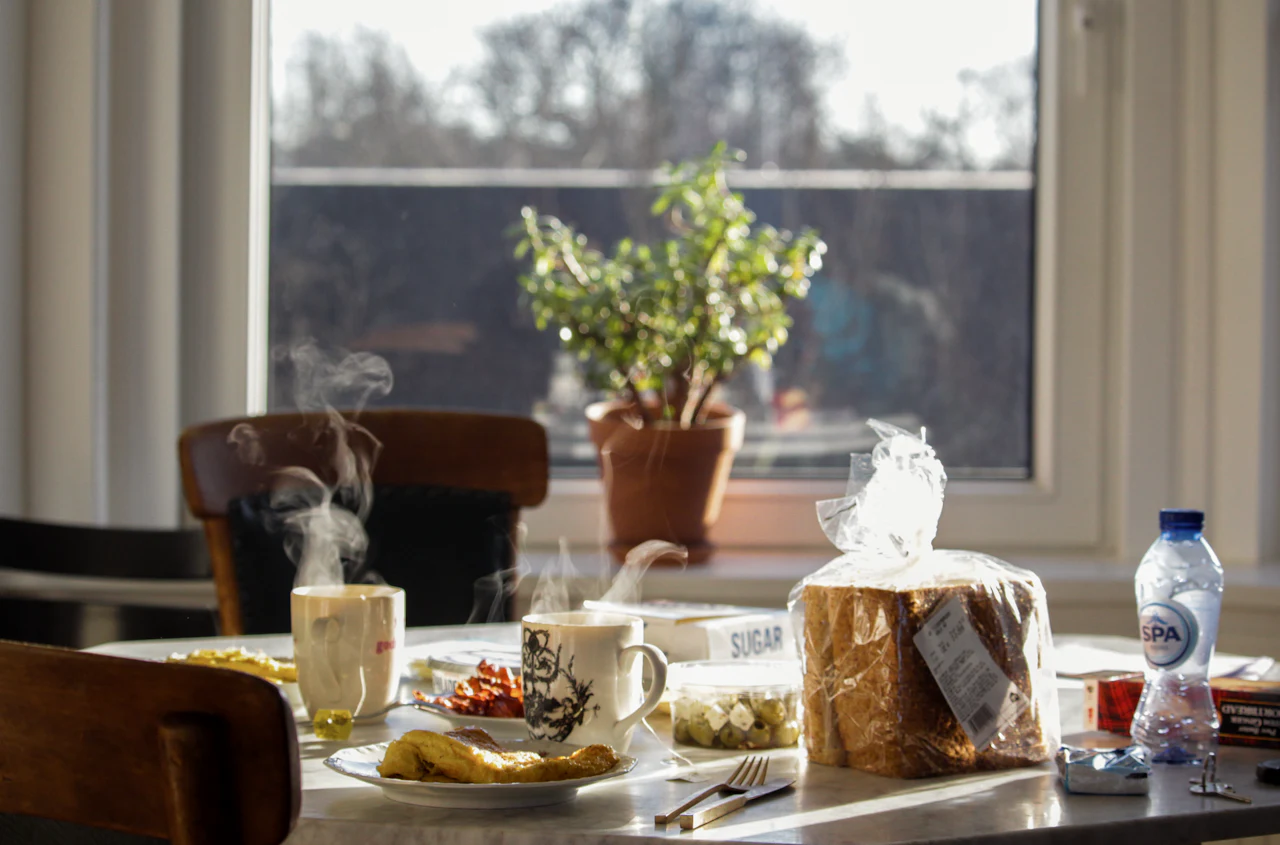 A breakfast table by a window, laid with coffee, bread and a potted plant
