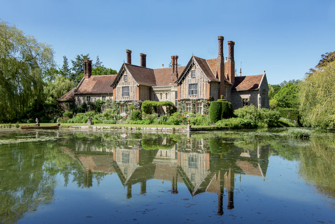 A timber-framed manor house with tall brick chimneys, reflected in still water beneath a blue sky