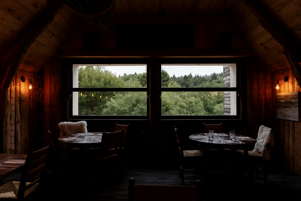 A timber-lined dining room with lamps, set tables and windows looking out to trees